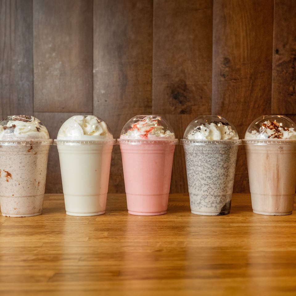Five colorful milkshakes topped with whipped cream sit on a wooden table against a rustic wood-paneled wall.