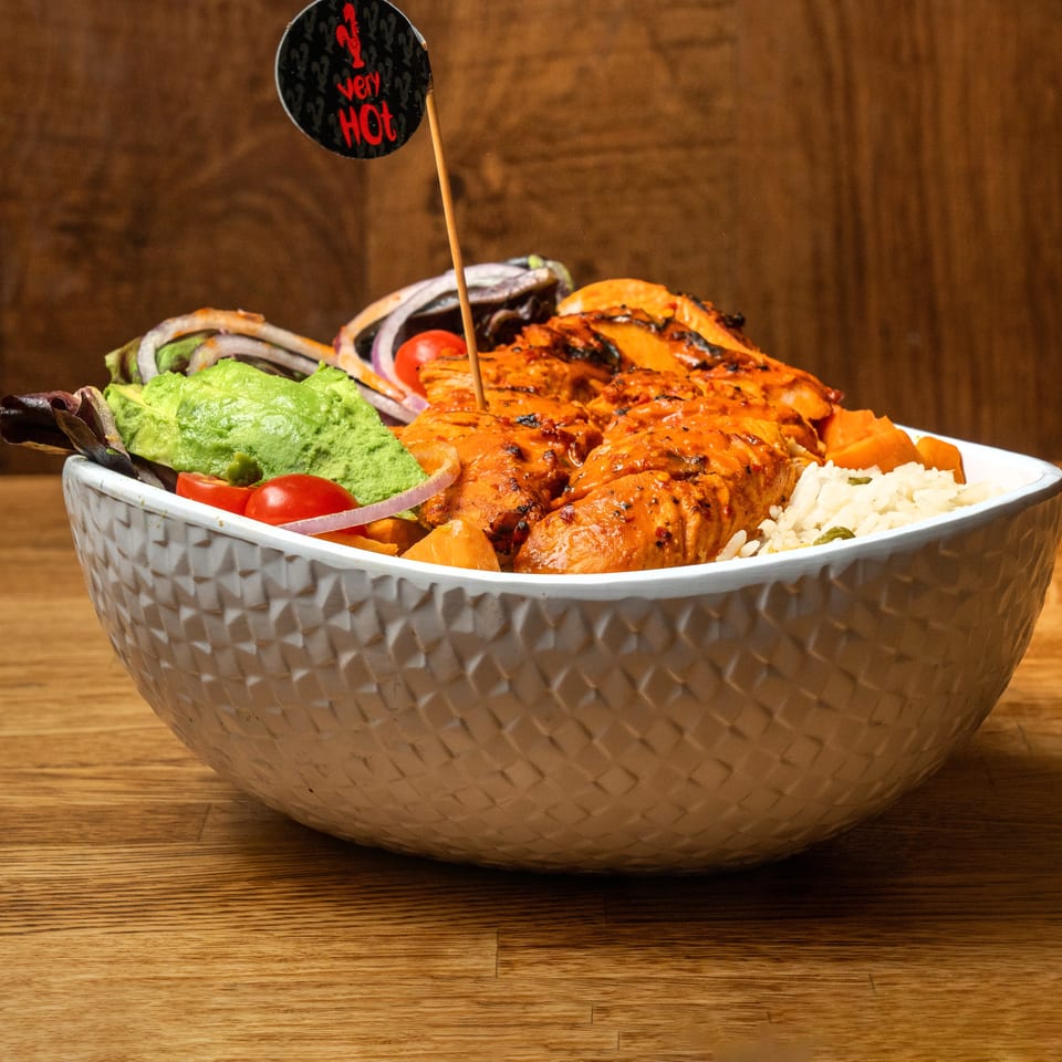 Grilled salmon bowl with white rice, guacamole, mixed greens, tomatoes, onions, and carrots on a wooden table.