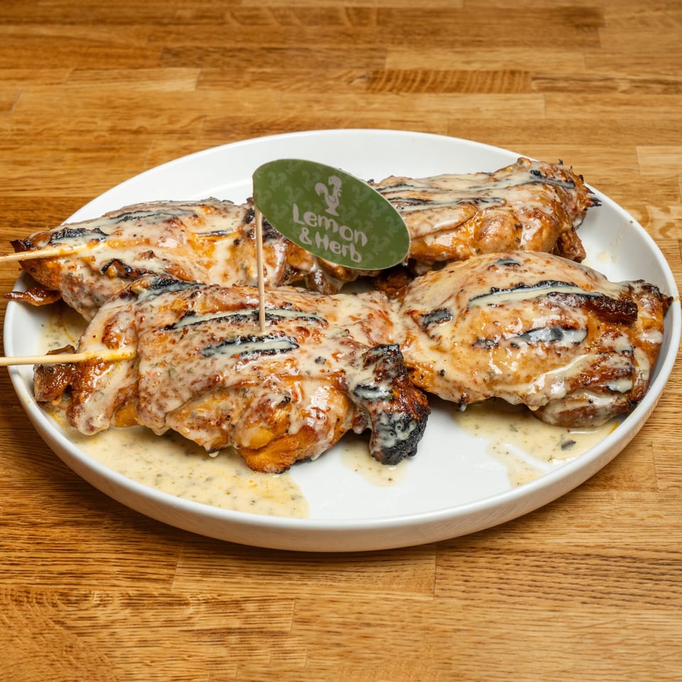 Grilled chicken skewers topped with creamy sauce on a white plate, garnished with a Lemon & Herb sign.
